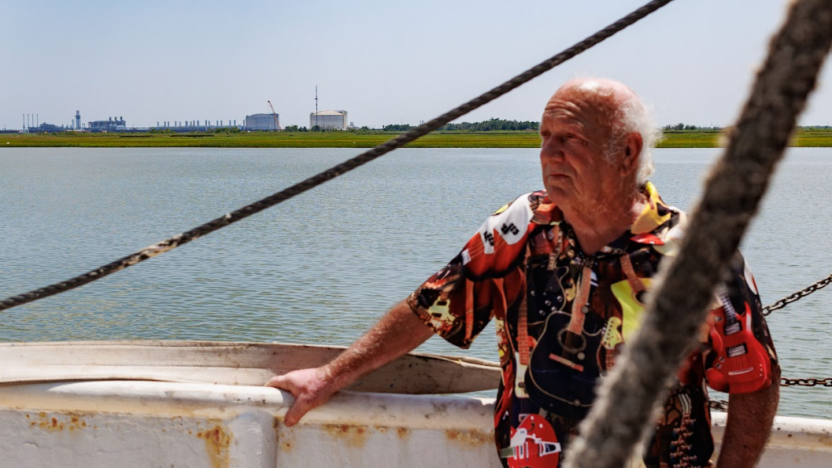 Senior mail in a colorful guitar print shirt, stands on his boat in the water, looking into the distance.