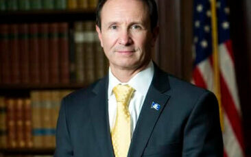 Louisiana governor, Jeff Landry, in jacket and tie before a bookcase and American flag.