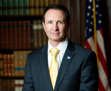 Louisiana governor, Jeff Landry, in jacket and tie before a bookcase and American flag.